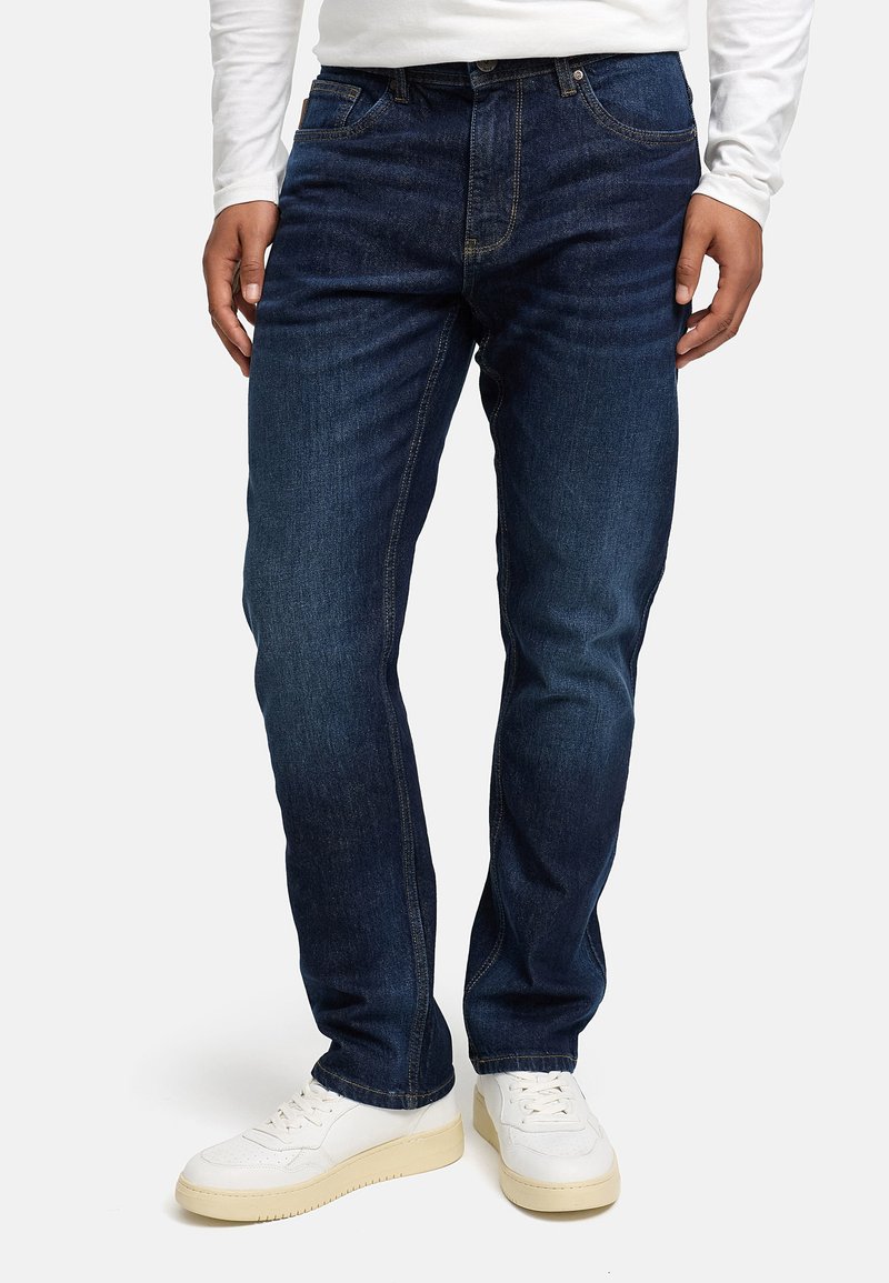 Dunkelblaue Denim-Jeans im Straight-Fit-Design, mit fünf Taschen und sandfarbenen Nähten. Getragen mit weißen Sneakers.
