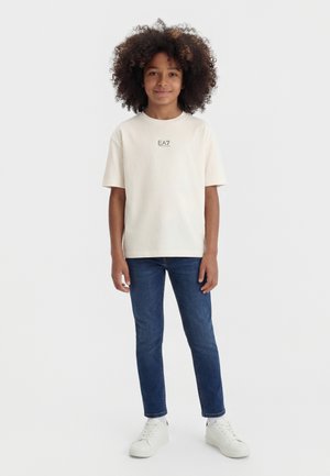 Dítě s kudrnatými vlasy stojí, má na sobě krémové tričko EA7, modré džíny a bílé tenisky, usmívá se na jednoduchém světlém pozadí.