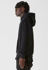 Melna kapuce ar priekšējo kabatu, novilcējamiem aukliņām un ribinātām piedurknēm. Zem tās redzama melna t-krekla virsma ar brīvu piegriezumu.