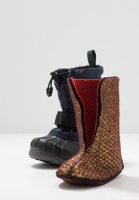 Zwei Stiefel: ein hoher, navyblauer wasserdichter Stiefel mit verstellbarem Kordelzug und schwarzer Sohle sowie ein kurzer, rot-gelb gemusterter Filzstiefel mit einer Naht.