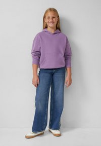 Světle fialová mikina s kapucí a přední kapsou, kombinovaná s širokými modrými džíny se bílými pruhy po stranách a krémovými teniskami.