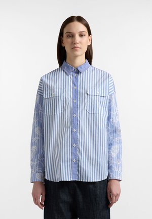 Camicia a righe blu e bianche con due tasche frontali, colletto con bottoni e motivi floreali decorativi sulla manica sinistra.