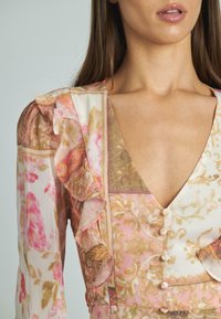 Kwiatowa sukienka z falbankami na rękawach w pastelowym różu, kremie i złocie. Charakteryzuje się głębokim dekoltem w serek oraz guzikami z przodu. Wykonana z lekkiego materiału.