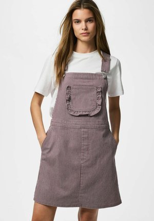 Giovane donna che indossa una t-shirt bianca e un vestito salopette a righe viola con tasca anteriore arricciata, mani nelle tasche laterali, sfondo neutro.