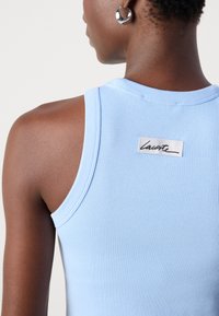 Débardeur côtelé bleu clair avec un col rond et un petit logo blanc. Il présente une texture lisse et un design sans manches.