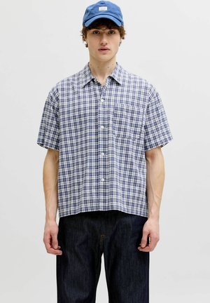 Jeune homme portant une chemise à carreaux bleue à manches courtes boutonnée, un jean foncé et une casquette de baseball bleue, debout devant un fond clair uni.