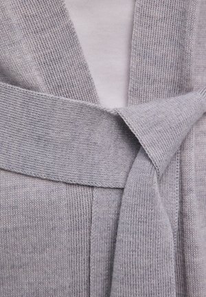 Hellgraue Strickjacke mit breitem Gürtel, locker zu einem Knoten gebunden, über einem weißen Unterhemd.