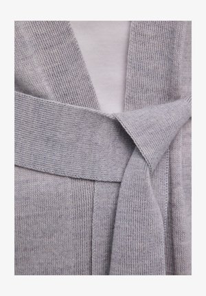 Hellgraue Strickjacke mit breitem Gürtel, locker zu einem Knoten gebunden, über einem weißen Unterhemd.