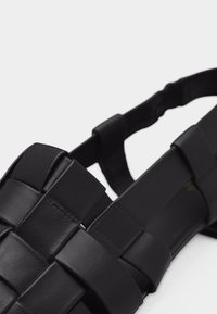 Κοντινό πλάνο μαύρου δερμάτινου σανδαλιού με φαρδιές πλεκτές λουρίδες και ελαστικό πίσω λουρί σε λευκό φόντο.