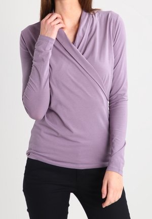 Frau trägt ein langärmliges, lavendelfarbenes Wickeloberteil und schwarze Hosen, posiert mit einer Hand am Hals und der anderen an der Seite.