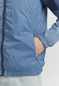 Giacca zip-up di colore azzurro chiaro, realizzata in materiale sintetico liscio. Presenta polsini elasticizzati e tasche laterali. Accento di colore blu marino in contrasto sulle spalle.