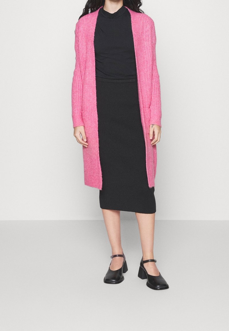 En kvinna i en lång rosa kofta över en svart klänning och svarta Mary Jane-skor, stående på ett ljusgrått golv mot en vit vägg.