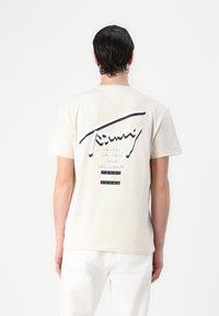 T-shirt en coton beige avec un graphique noir courbé et du texte au dos, comportant des noms de villes et "TOMMY JEANS" dans des carrés noirs.