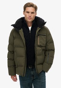 Olijfgroene donzen jas met een grote zwarte kraag, ritszakken en het "Superdry"-logo. Draagt een marineblauwe kabelgebreide trui eronder.