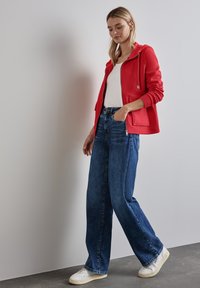 Felpa rossa con zip sopra una t-shirt bianca, abbinata a jeans blu a gamba larga e sneakers bianche. Texture lisce con dettagli di design semplici.