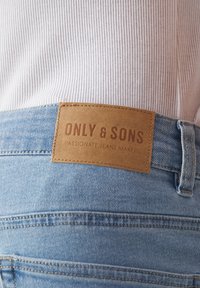 Vaaleansiniset farkut, joissa on ruskea nahkapaikka, jossa lukee "ONLY & SONS" ja "PASSIONATE JEANS MAKERS". Niissä näkyy niittejä ja ompelutarkkuutta.
