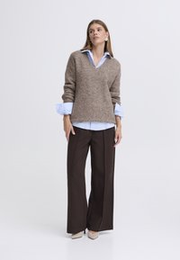 Rjav pulover iz volnene mešanice z V-izrezom, oblečen čez svetlo modro srajco z gumbi. Rjave široke hlače in bež čevlji z ostrim prstom.