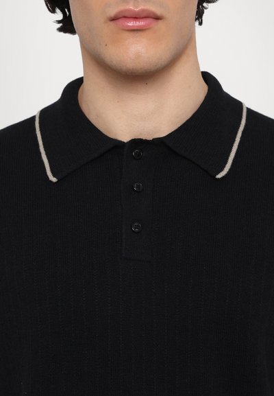 Polo shirt noir en tricot avec une texture côtelée, doté d'un col beige contrastant et de trois boutons à l'avant pour la fermeture.