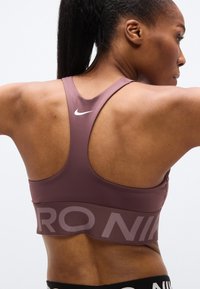 Αθλητικό σουτιέν Nike σε μωβ απόχρωση με σχεδιασμό racerback. Διαθέτει φαρδιά ζώνη με το λογότυπο "PRO". Ομαλή υφή και κατασκευή χωρίς ραφές.