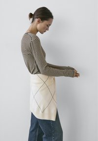 Gestreiftes Langarmoberteil in Braun und Creme, kombiniert mit einem cremefarbenen Strickpullover mit Rautenmuster und dunkelblauen Jeans mit weitem Bein.
