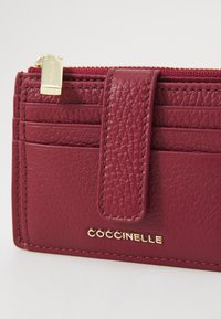 Portafoglio in pelle bordeaux con superficie texturizzata, hardware dorato, numerosi scomparti per carte e una tasca con cerniera. Nome del brand "Coccinelle" impresso.