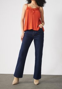 Top a maniche a canottiera plissettato arancione con dettagli in pizzo, abbinato a jeans a gamba larga blu navy e décolleté beige, il tutto su uno sfondo neutro.