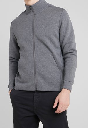 Mann trägt eine graue Jacke mit Reißverschluss und seitlichen Taschen sowie eine schwarze Hose, steht mit einer Hand in der Tasche vor einem schlichten Hintergrund.