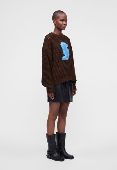 Personne debout portant un pull marron en tricot avec un motif hippocampe bleu, une jupe en cuir noire et des bottes noires mi-mollet sur un fond uni.