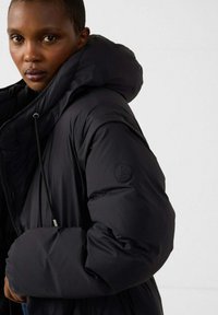 Schwarze Steppjacke mit großer Kapuze, die ein kreisrundes Logo auf dem Ärmel, eine strukturierte Stoffoberfläche und einen Kordelzug am Halsausschnitt aufweist.