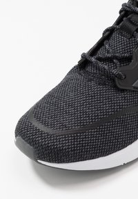 Texturovaná černá sportovní bota s síťovinovým svrškem, syntetickými akcenty, zaoblenou špičkou a bílou podrážkou. Obsahuje černé tkaničky a brandingový hardware.