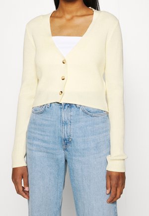Femme portant un cardigan court jaune clair avec trois boutons sur un top blanc, associé à un jean taille haute bleu clair.