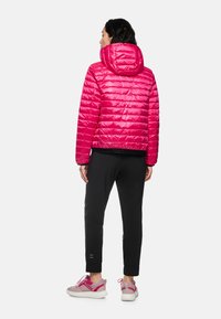 Woman facing away wearing a bright pink quilted hooded jacket, black tapered pants, and pink and white sneakers against a plain background.