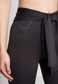 Leggings neri a vita alta con un laccio in tessuto in vita, tasca laterale e texture liscia, caratterizzati da dettagli di cucitura a contrasto.
