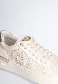 Beige sneakers med et struktureret mønster, guld accenter og cremefarvede snørebånd. Har en polstret krave og logo detaljer på siden.