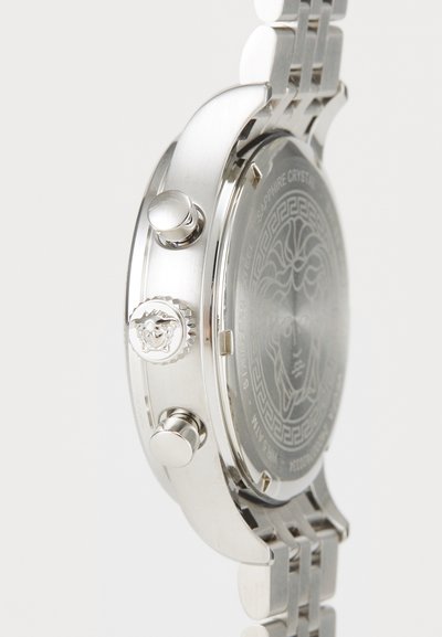 Silverfärgad rostfritt stål klocka med tre knappar, en texturerad krona med en medaljong och en mönstrad baksida.