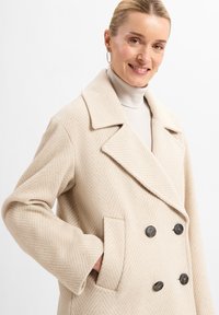 Beige visgraatjas met een dubbele rij knopen, grote revers en zwarte knopen. Bevat zijzakken en een gestructureerde stof.