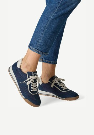 Füße tragen marineblaue und weiße Schnürsneaker mit braunen Sohlen, kombiniert mit blauen, gekürzten Jeans aus Denim vor einfarbigem Hintergrund.