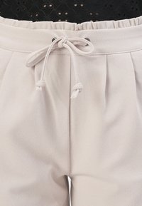 Pantalones de tejido suave color beige claro con cinturilla elástica y cordón ajustable. Presentan pliegues frontales y una textura suave.