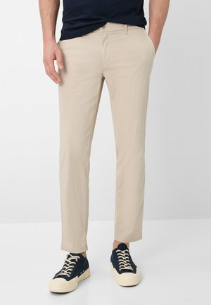 Mann trägt beige gerade geschnittene Hose mit seitlichen Taschen und schwarze Low-Top-Sneaker mit weißen Sohlen und Schnürsenkeln, steht vor weißem Hintergrund.