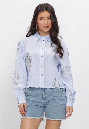 Vrouw met lang donker haar draagt een lichtblauw gestreept bloesje met zilveren bloemenpatronen en lichte denimshorts, staand tegen een witte achtergrond.