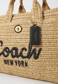 Sac en toile tissée en paille avec un grand logo noir brodé, fermeture à bouton doré et une étiquette en cuir noir avec un accent en chaîne.