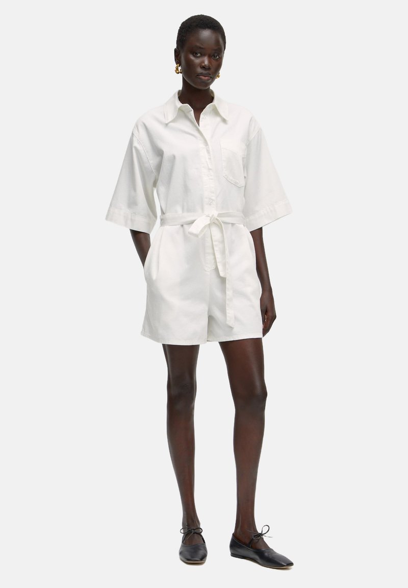 Witte katoenen jurk met korte mouwen en een strik taille, met een patchzak en een ontspannen pasvorm, gecombineerd met zwarte platte schoenen.