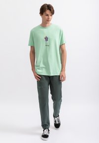 T-shirt en coton vert clair avec un design graphique coloré ; associé à un pantalon de jogging vert foncé et des baskets noires avec des lacets blancs.