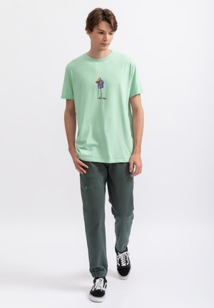 T-shirt en coton vert clair avec un design graphique coloré ; associé à un pantalon de jogging vert foncé et des baskets noires avec des lacets blancs.