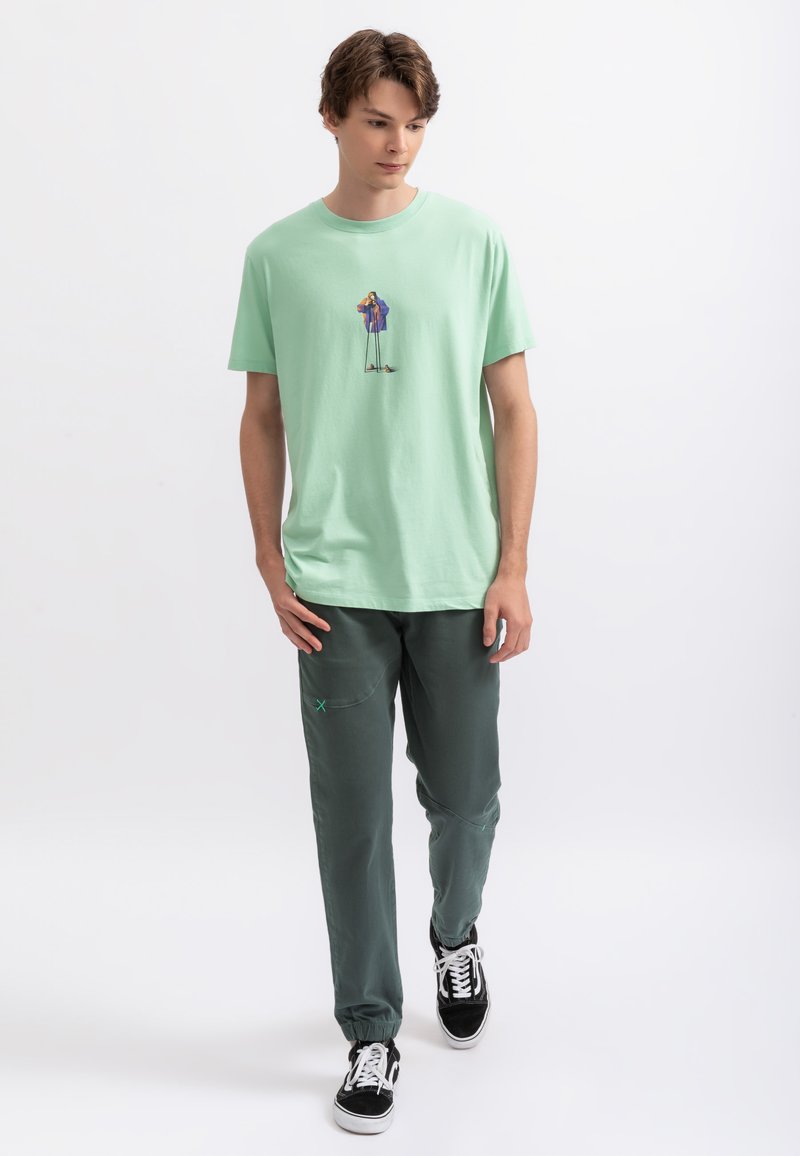 T-shirt en coton vert clair avec un design graphique coloré ; associé à un pantalon de jogging vert foncé et des baskets noires avec des lacets blancs.