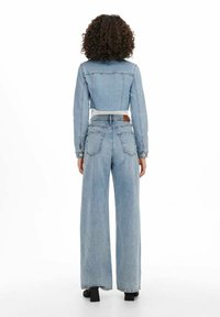 Vaaleansininen farkkutakki, jossa on lyhyt malli ja raakareuna, yhdistettynä leveälahkeisiin vaaleansinisiin farkkuihin. Takaa näkyy taskut ja vyötärön yksityiskohdat.