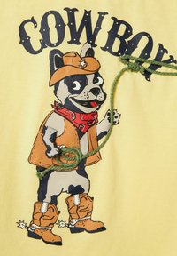 Rysunkowy pies w żółtej koszuli; nosi brązowy kapelusz cowboya, czerwony bandana, beżową kamizelkę i pomarańczowe buty; zielony detal ze sznurkiem. Tekst brzmi "COWBOY."