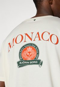 Biała koszulka z dużym czerwonym napisem "MONACO" oraz zielonym emblematem z koroną i rakietami tenisowymi, wykonana z miękkiej bawełny, luźny krój.