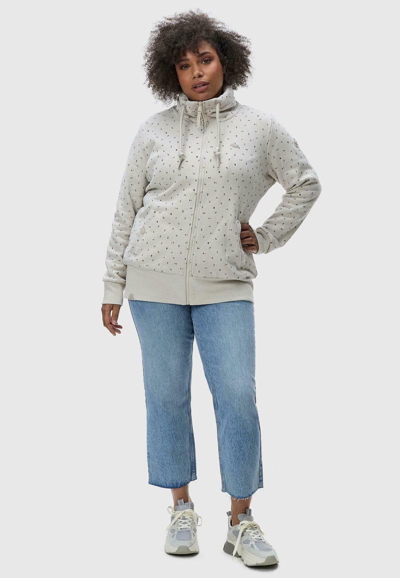 Lichtgrijze zip-up hoodie met een hoge kraag, versierd met een verspreid patroon van kleine gekleurde stippen, gecombineerd met blauwe cropped jeans en sneakers.