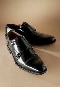 Zwarte leren dresschoenen hebben een gepolijste afwerking, dubbele gesps accentueringen en een getextureerde neuskap, tegen een neutrale achtergrond.
